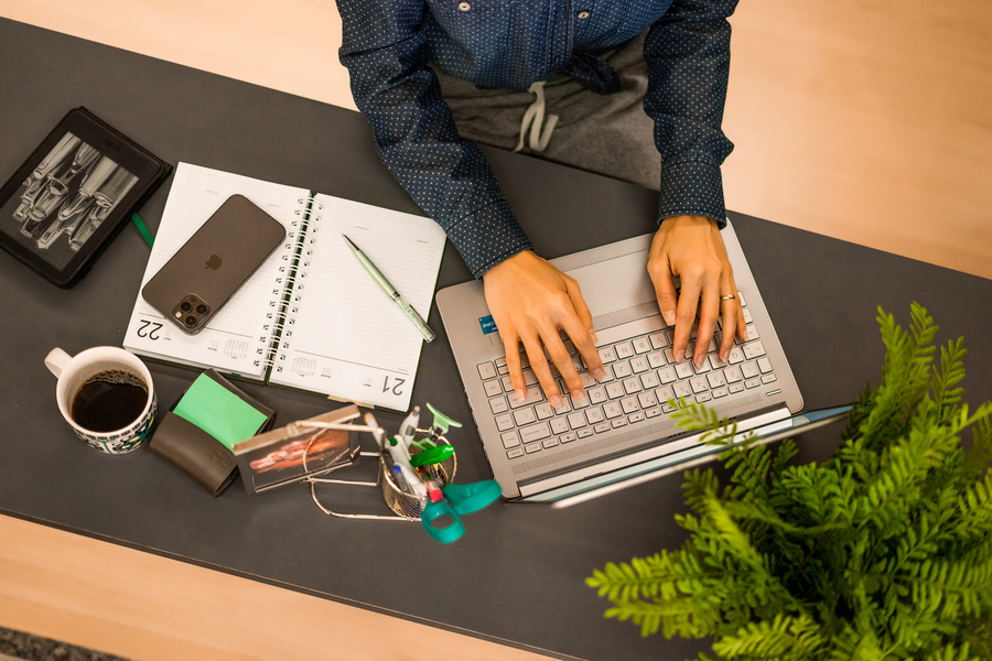 Pessoa trabalhando no computador ao lado de uma planta em apartamento com caderno e celular na mesa